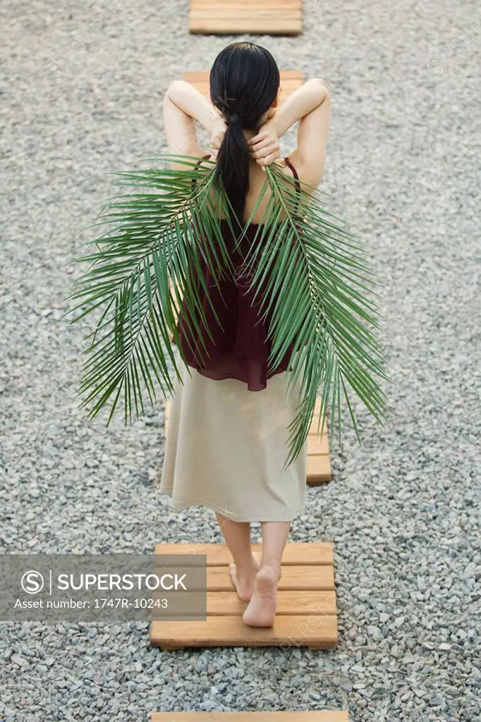 Woman walking on footpath, holding palm leaves behind back