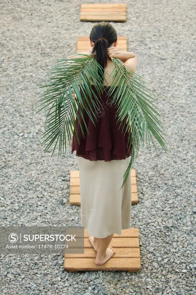 Woman standing on foothpath, carrying palm leaves behind back, rear view