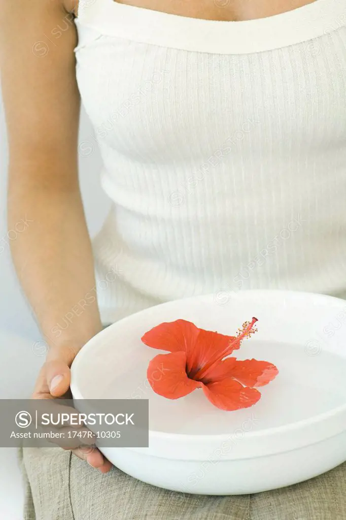 Woman holding hibiscus blossom in bowl of water on lap, cropped view