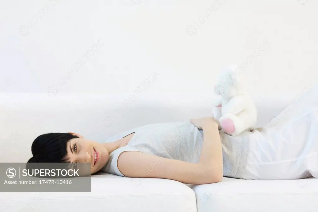 Woman lying on back on sofa, teddy bear sitting on her stomach, smiling at camera