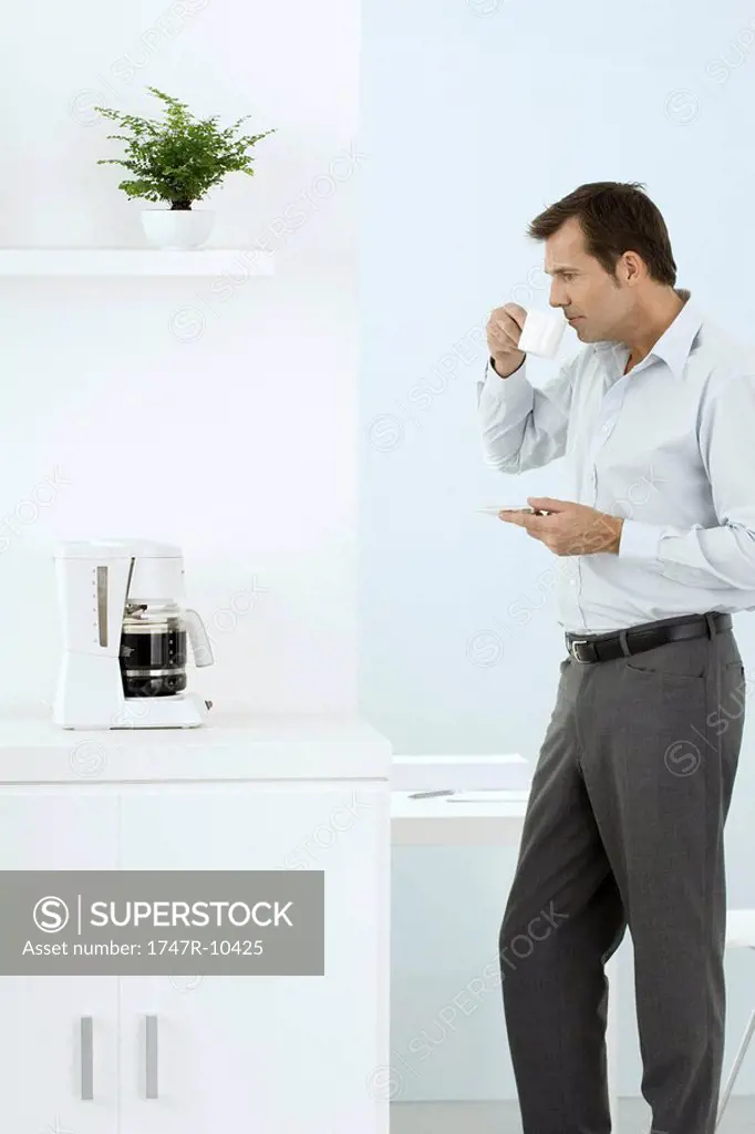 Man standing by coffee maker, taking a sip of coffee, holding saucer