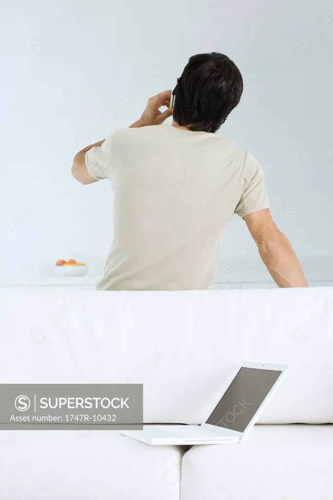 Man talking on cell phone, leaning against sofa with laptop on it, rear view