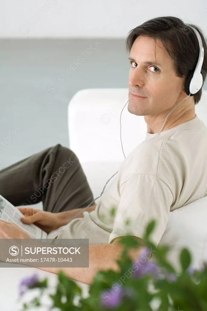 Man listening to headphones, looking over shoulder at camera, holding newspaper