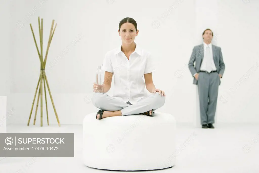 Professional woman sitting on ottoman, holding water bottle, businessman and bamboo in background
