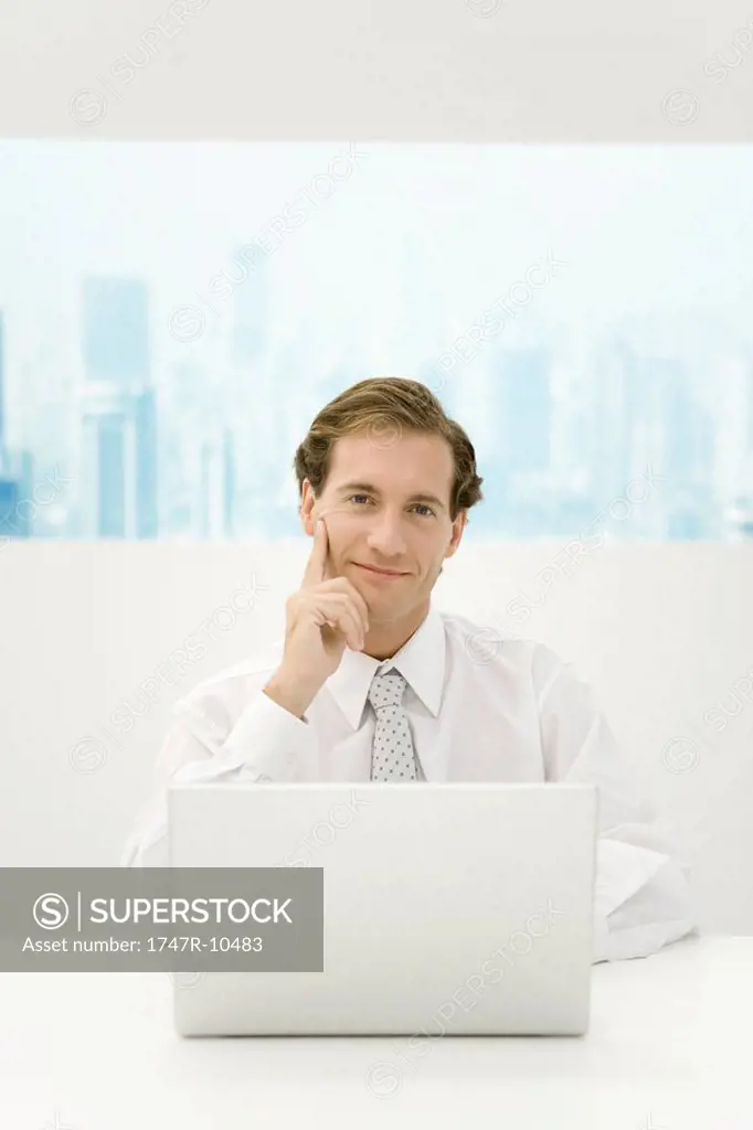 Businessman sitting with laptop computer, holding head, smiling at camera