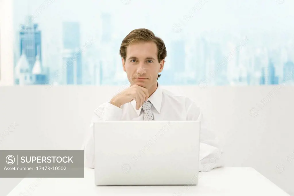Businessman sitting in front of laptop computer, hand under chin, looking at camera
