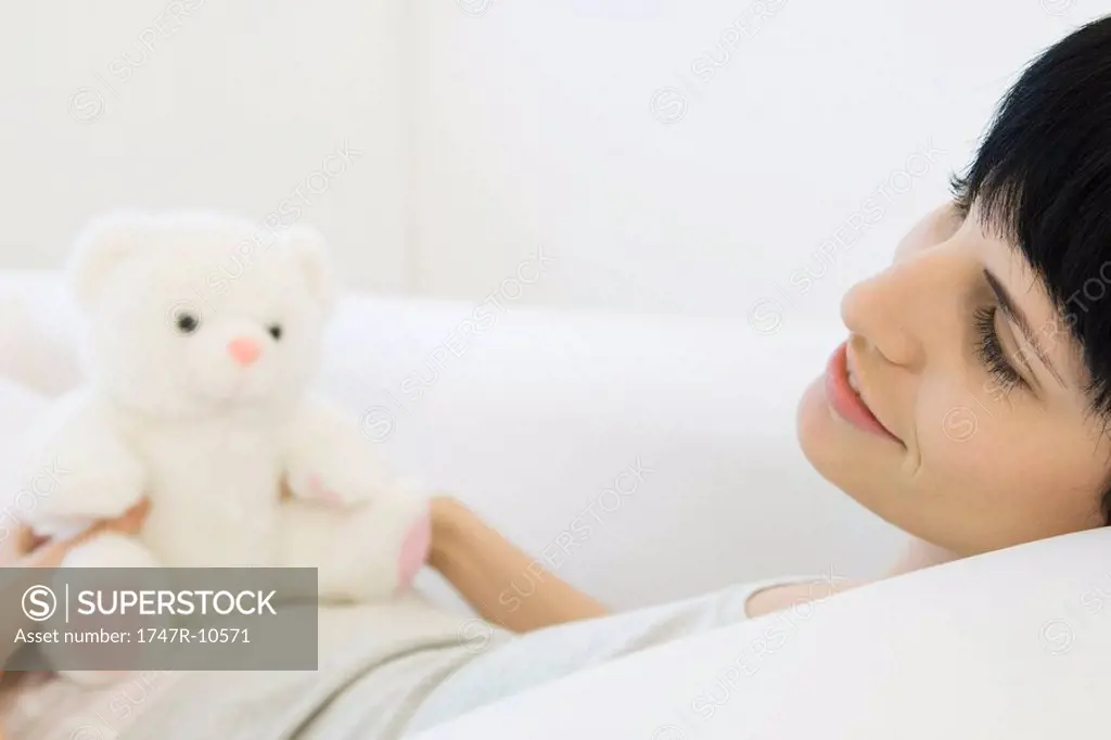 Woman lying on back on sofa, teddy bear sitting on her stomach, looking away