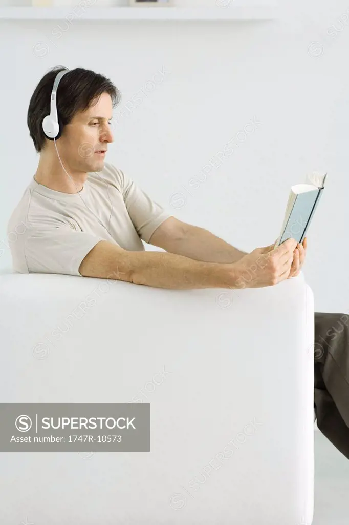 Mature man sitting in armchair, reading book and listening to headphones