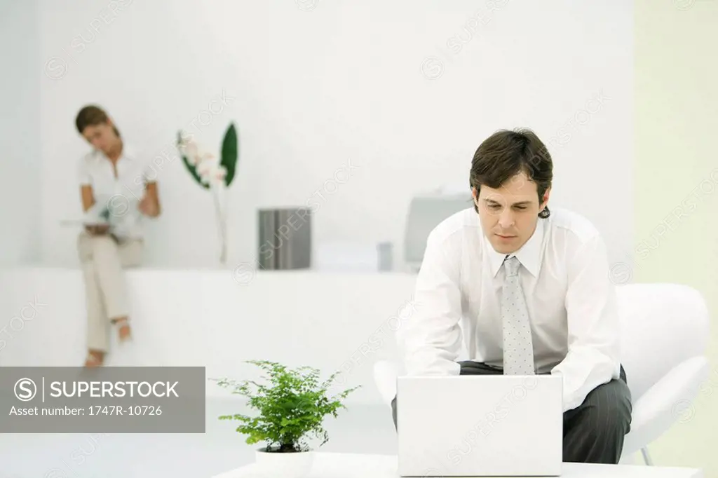 Professional man using laptop computer in casual office, female colleague reading in background