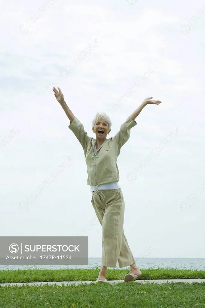 Senior woman standing outdoors in seaside park with arms raised, smiling happily
