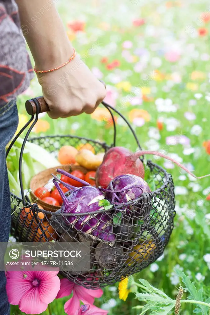 Person standing in field of wildflowers, carrying basket full of fresh produce, cropped view