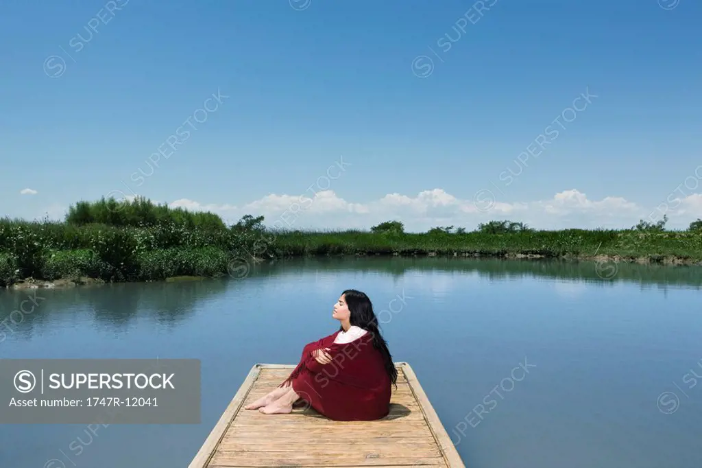 Woman sitting on dock with blanket wrapped around her, eyes closed