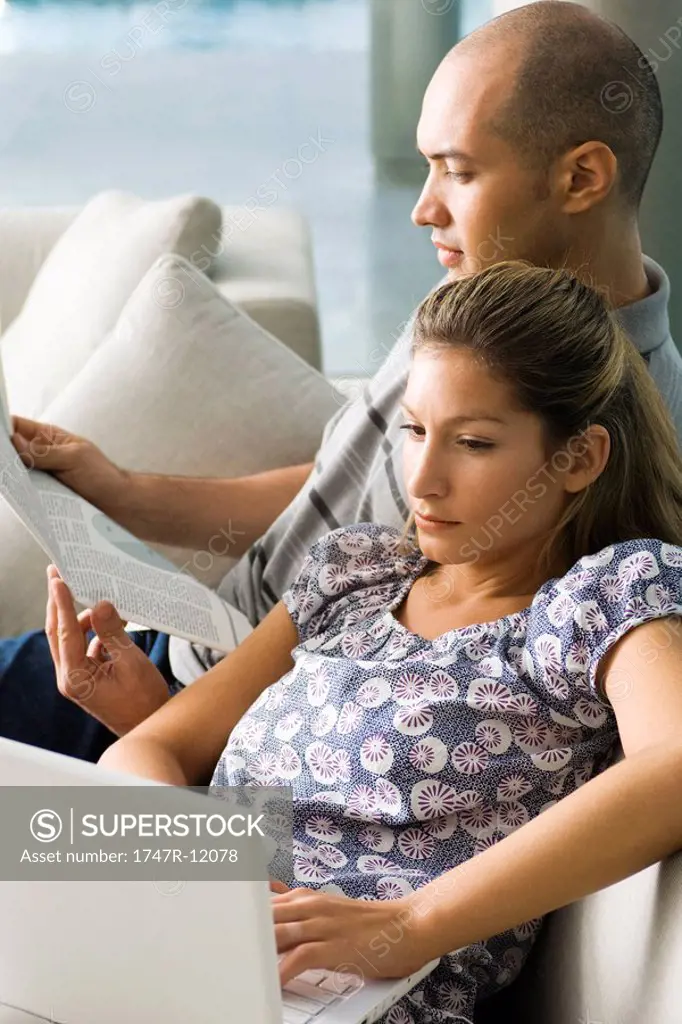 Couple relaxing together on sofa, man reading and woman using laptop computer