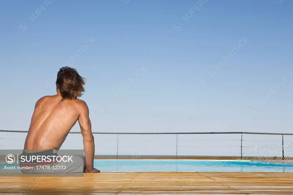 Man sitting poolside, dipping feet in water, rear view