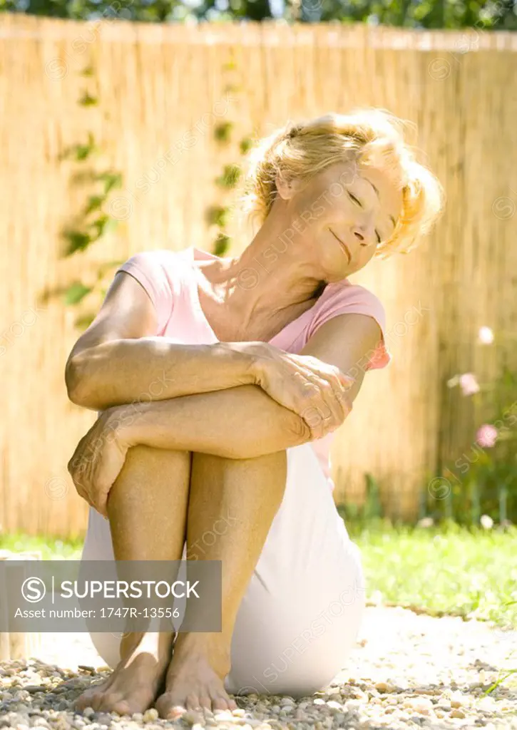 Senior woman sitting on ground outdoors, hugging knees, eyes closed, smiling