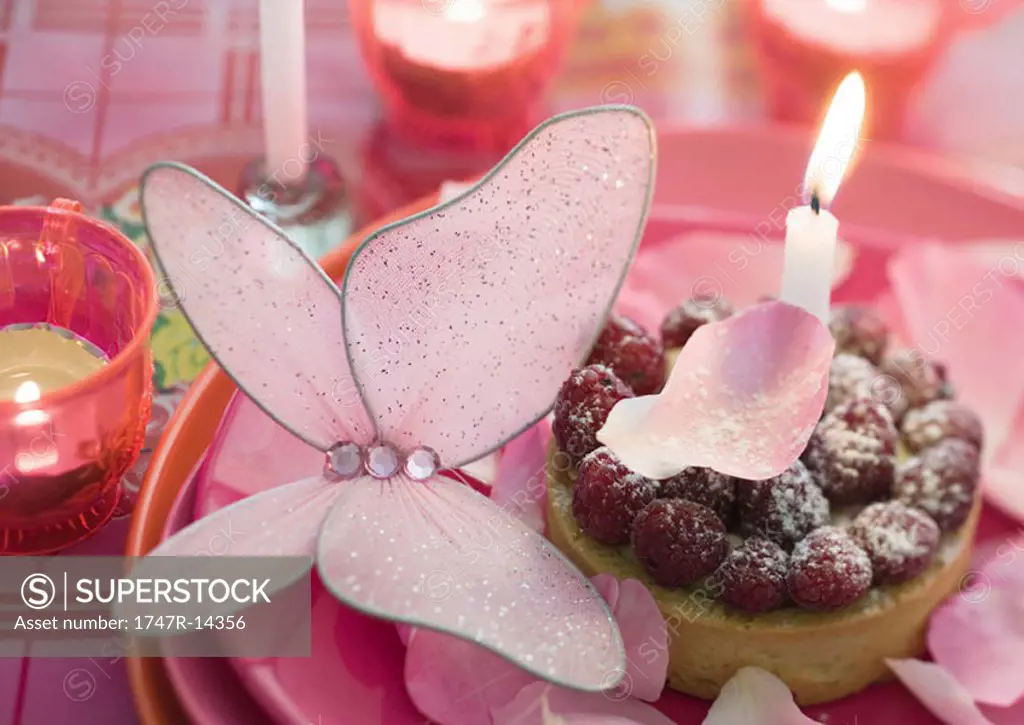 Raspberry tartlet on plate decorated with candle, butterfly and rose petals
