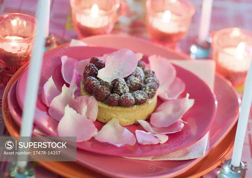 Raspberry tartlet on plate decorated with rose petals, surrounded with candles