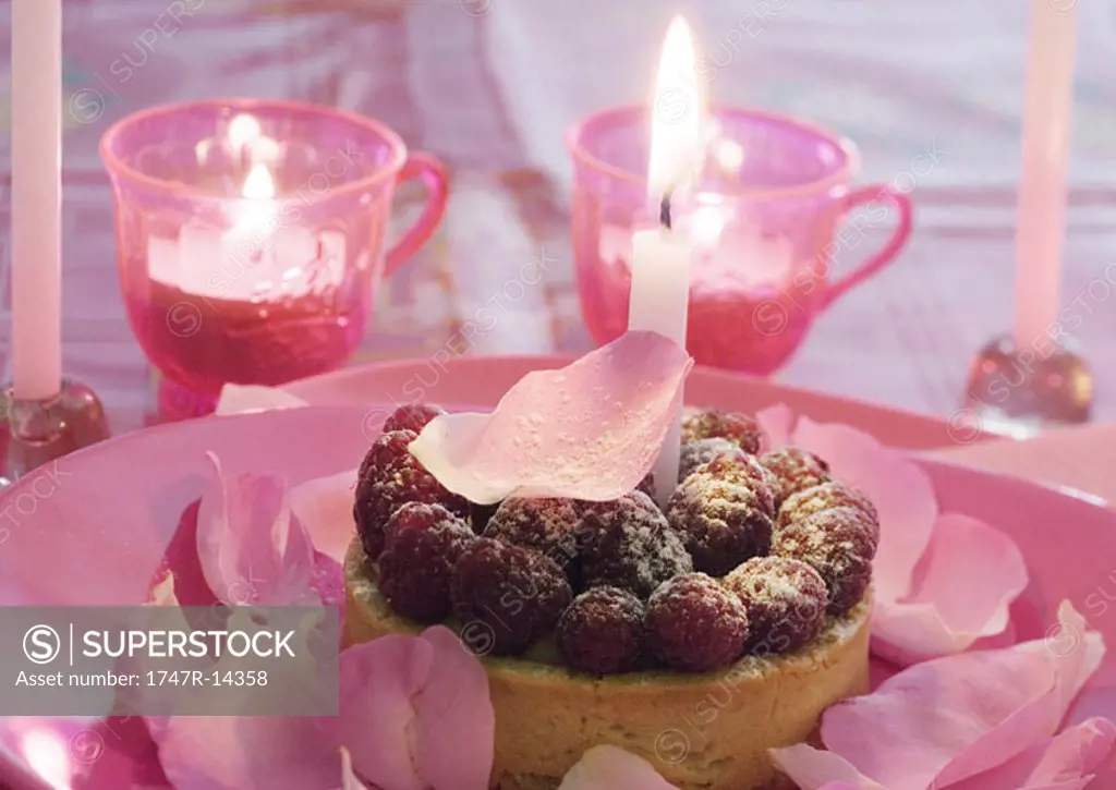 Raspberry tartlet on plate decorated with rose petals, surrounded with candles
