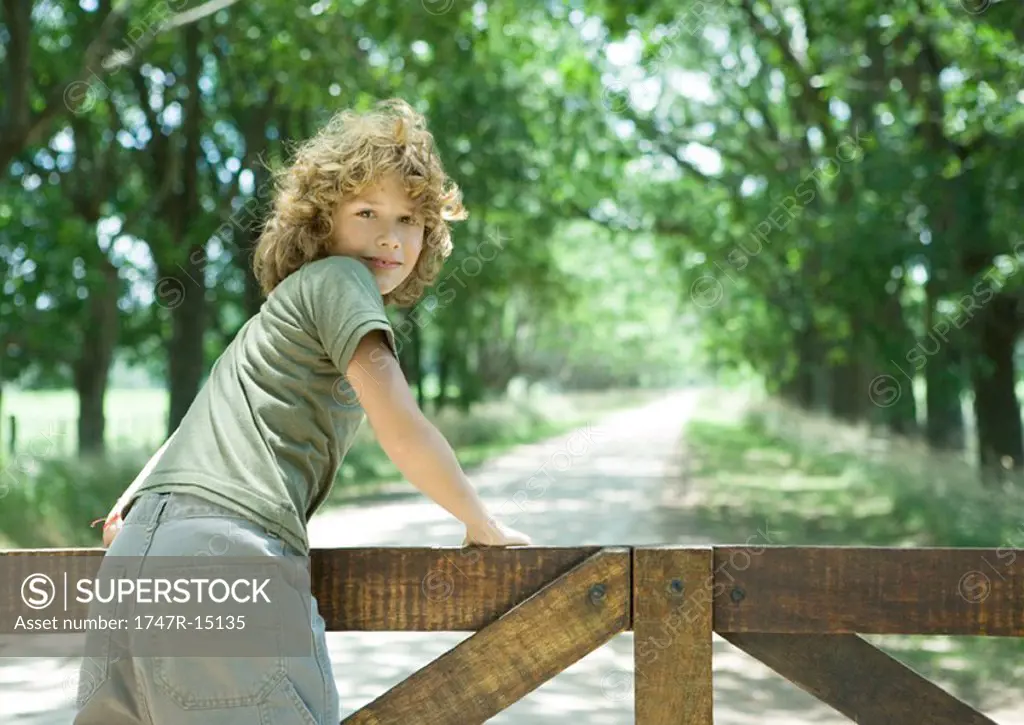 Boy leaning against wooden fence, looking over shoulder at camera