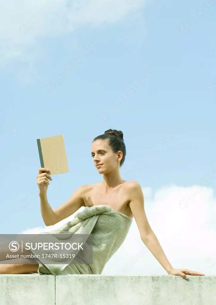 Woman sitting wrapped in towel, reading book, sky in background