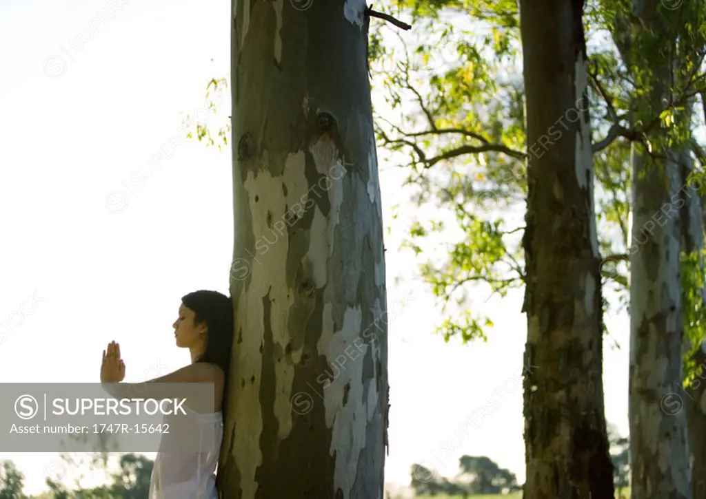 Woman standing with hands in prayer position, leaning against tree