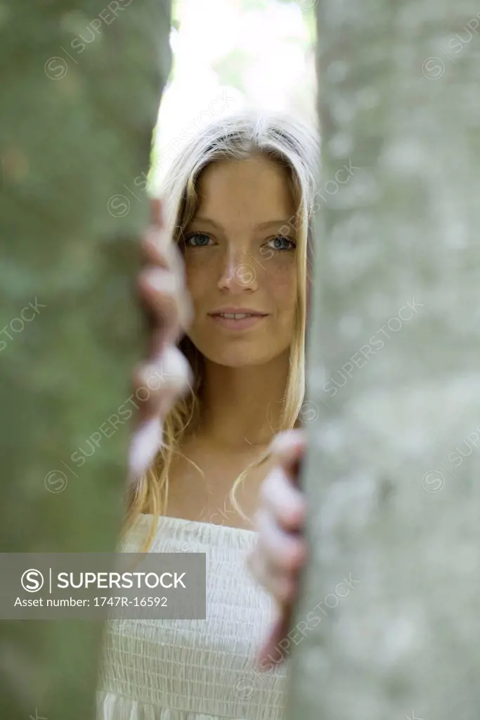 Young woman peering out from behind tree trunks
