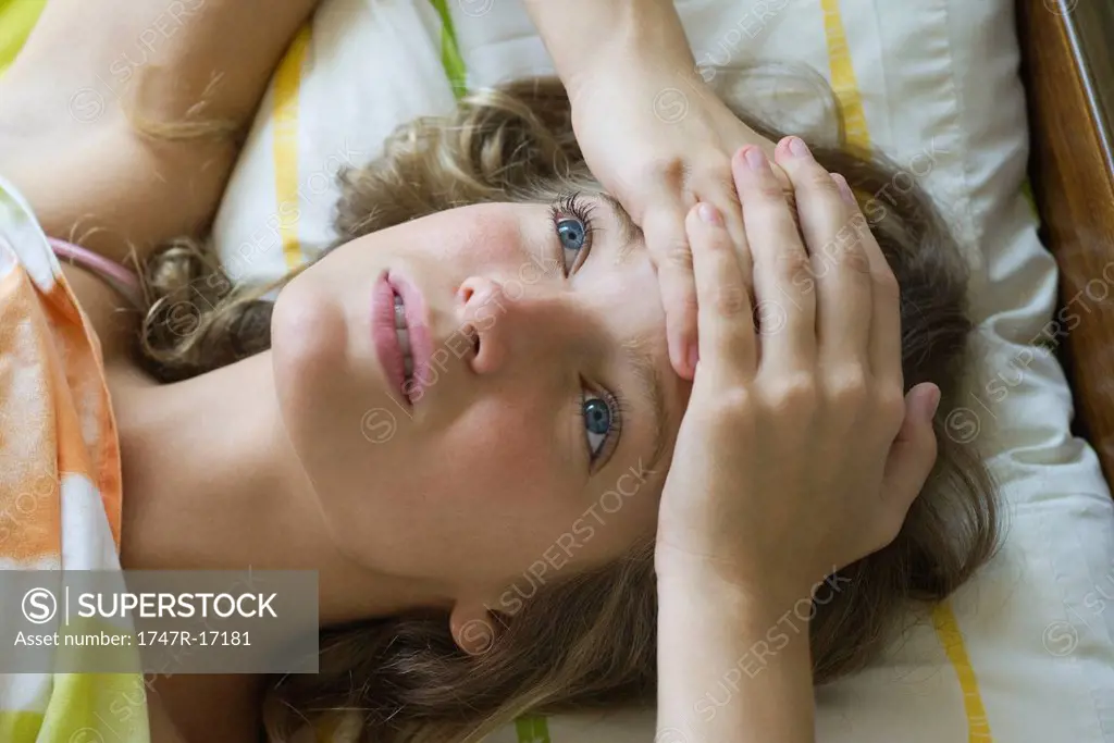 Young woman lying on bed with hands clasped on forehead, looking up in thought