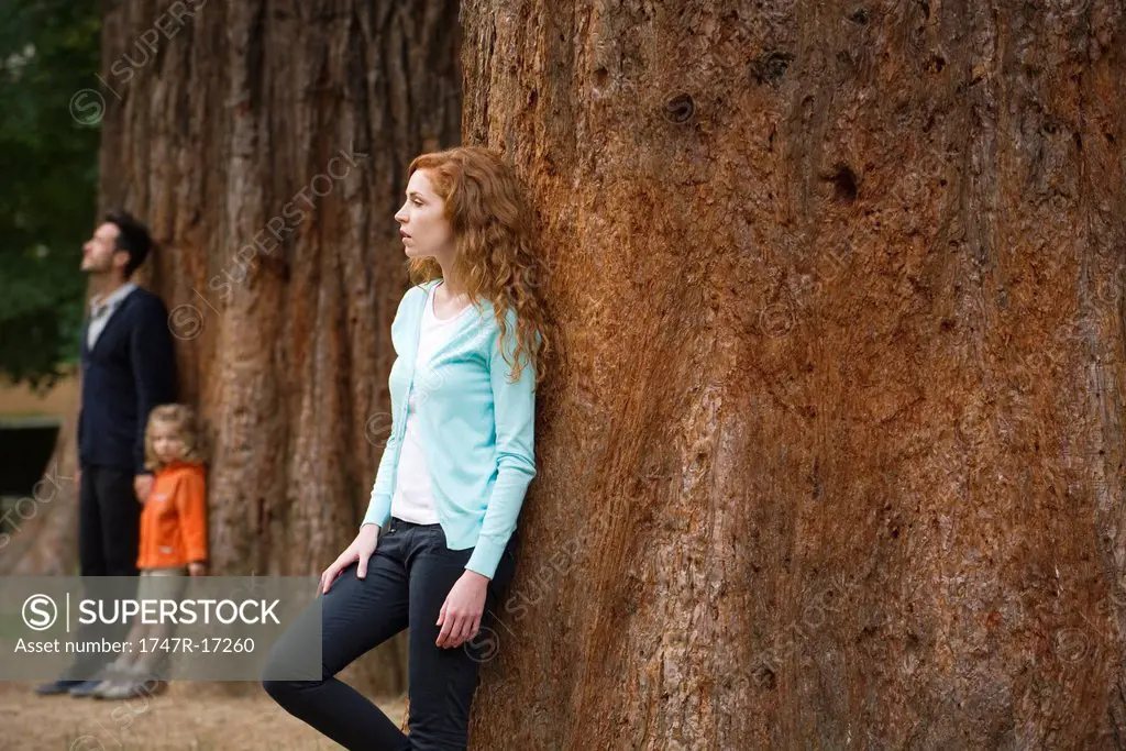 Mother leaning against tree trunk, husband and daughter separate in background