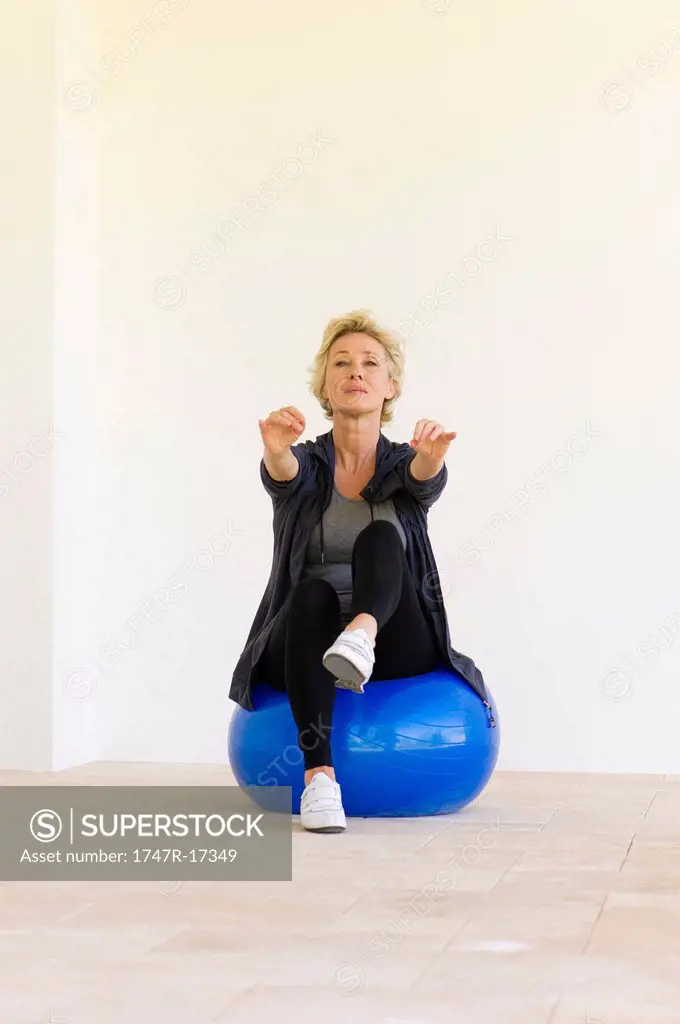 Mature woman sitting on fitness ball with arms out and one leg up