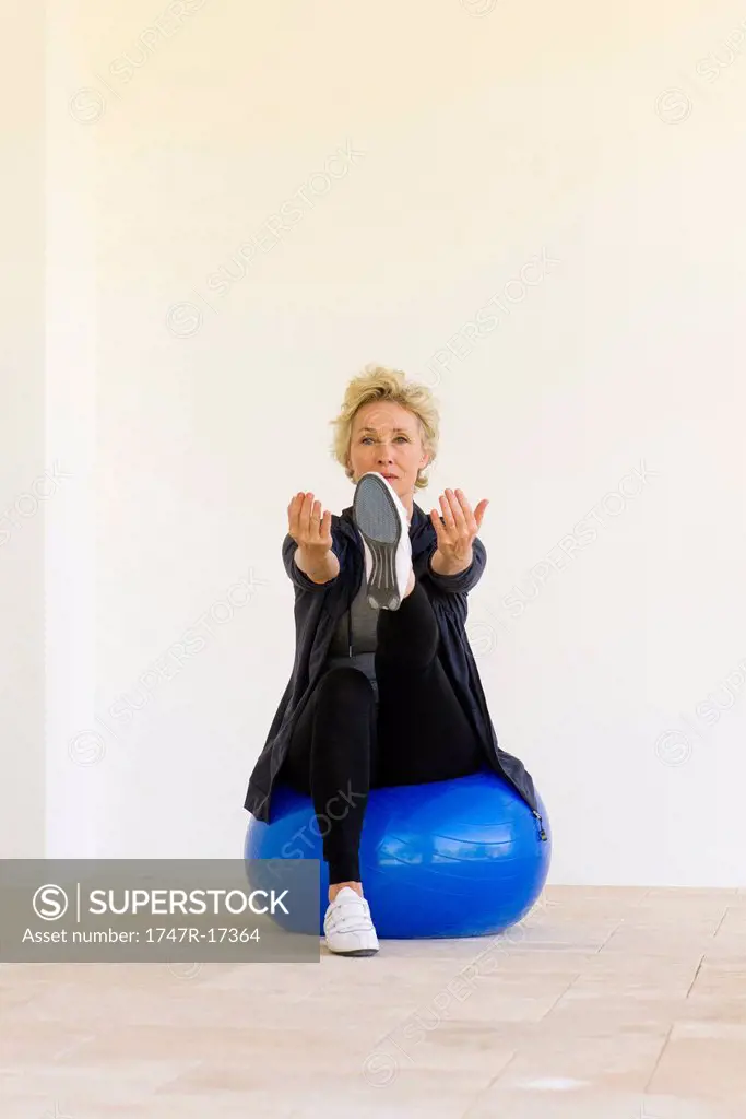 Mature woman sitting on fitness ball with arms out and one leg up