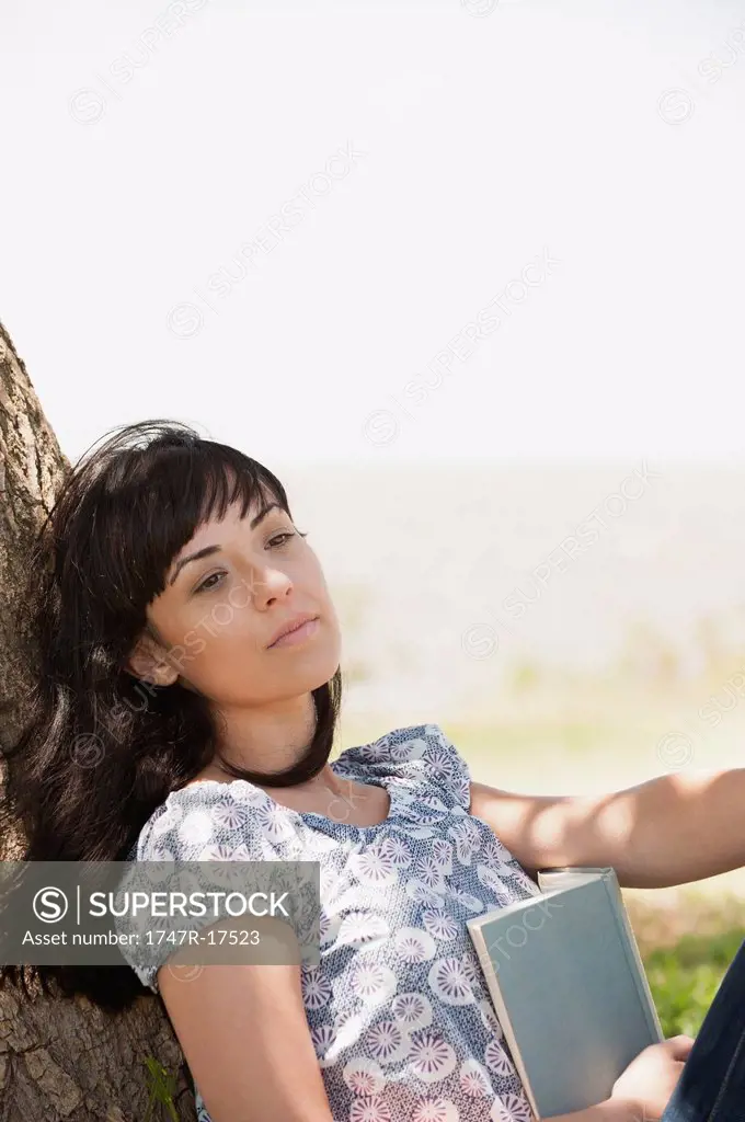 Young woman leaning against tree trunk holding book