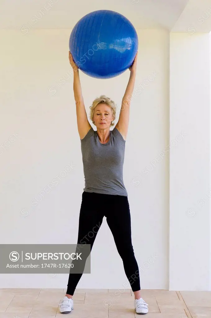 Mature woman holding fitness ball overhead with eyes closed