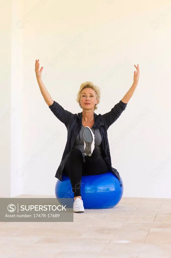Mature woman sitting on fitness ball with arms raised and one leg up