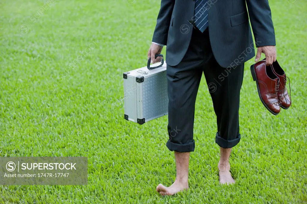 Businessman walking barefoot on grass carrying shoes and briefcase