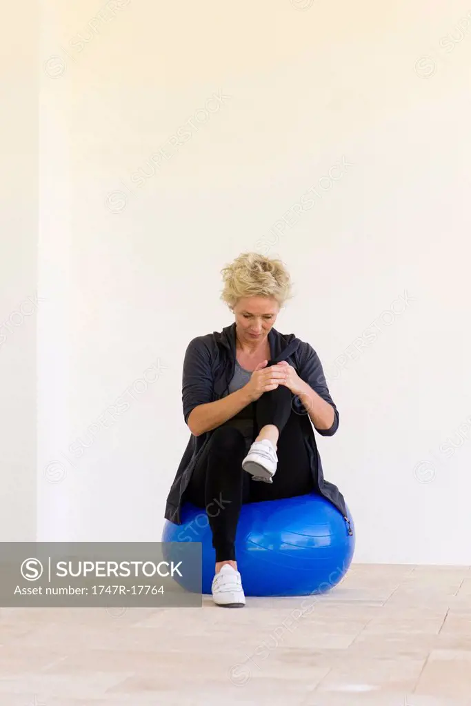 Mature woman sitting on fitness ball, hugging one knee