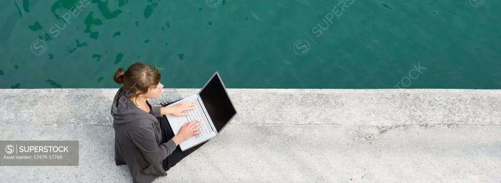 Young woman sitting beside canal, using laptop computer
