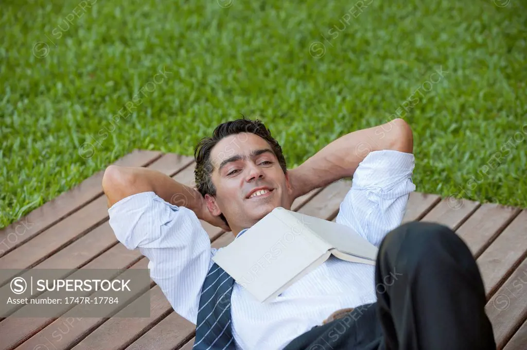 Mid_adult man lying outdoors with hands behind head and book on chest
