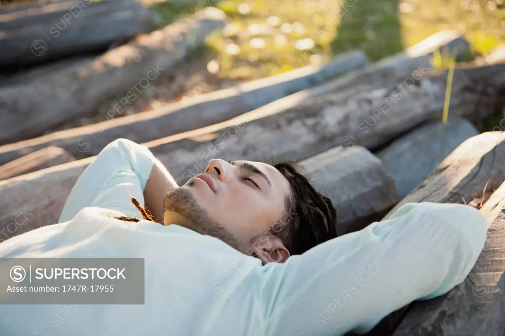 Young man lying on pile of logs, eyes closed