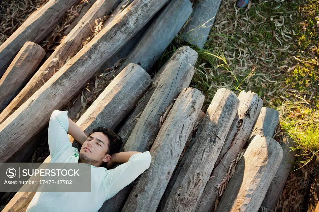 Young man lying on pile of logs, eyes closed