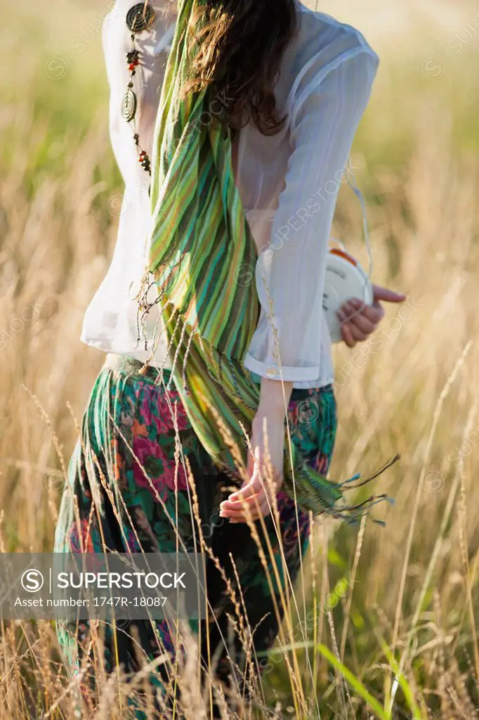 Young woman walking through field with portable CD player, rear view