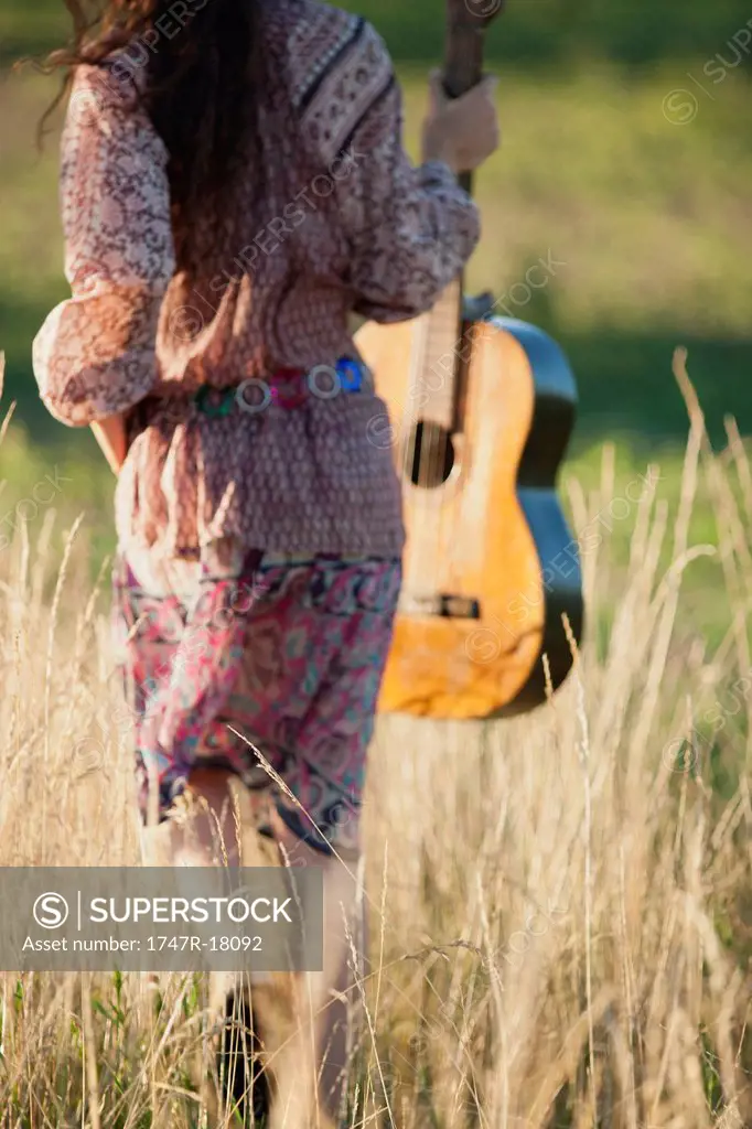 Woman walking through field with guitar, rear view