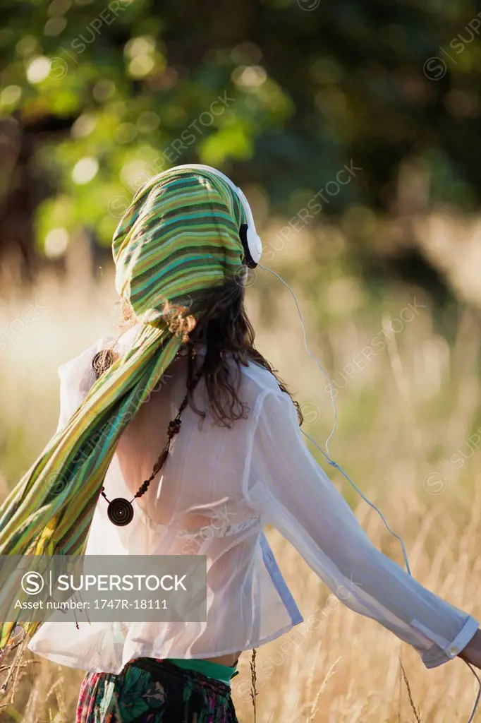 Woman walking through field, listening to headphones, rear view