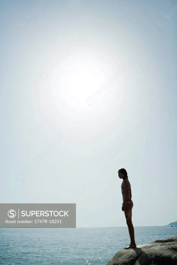 Girl in bikini standing on edge of cliff looking at ocean