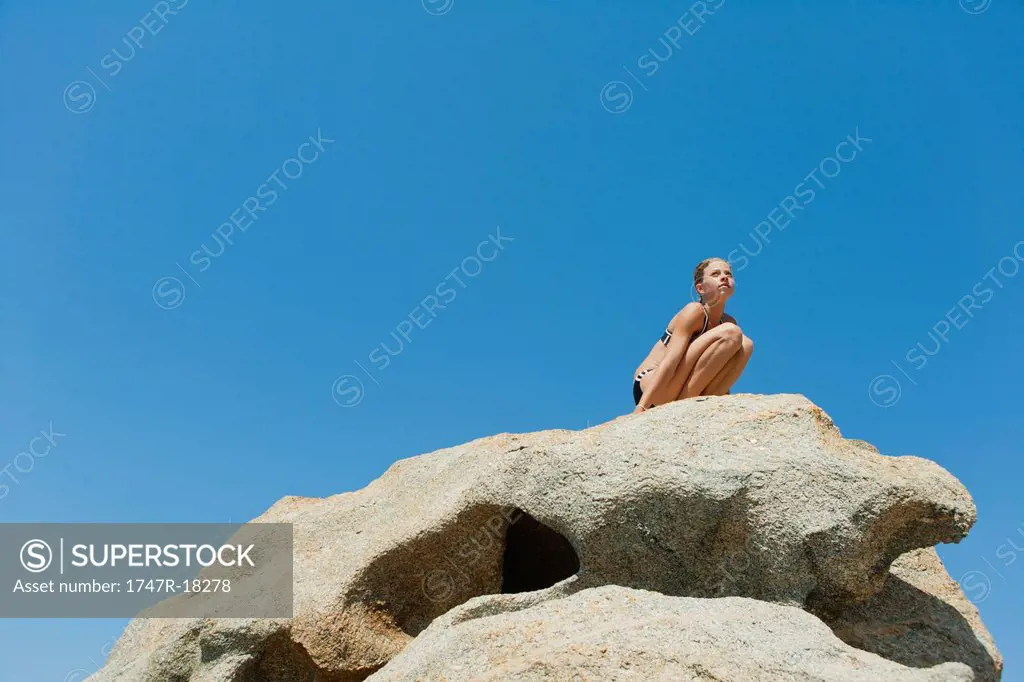 Preteen girl in bikini crouching on rock against blue sky, low angle view