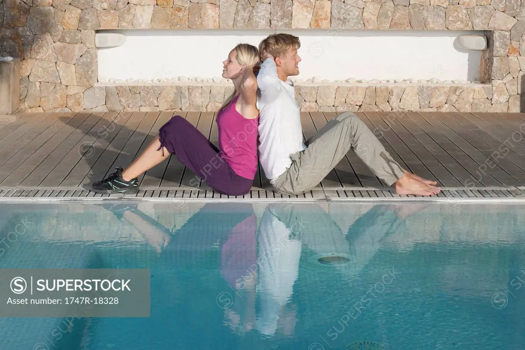 Young couple sitting back to back at poolside, hands behind heads