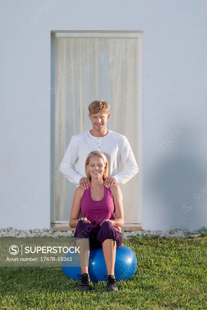 Young woman sitting on fitness ball, boyfriend giving her massage