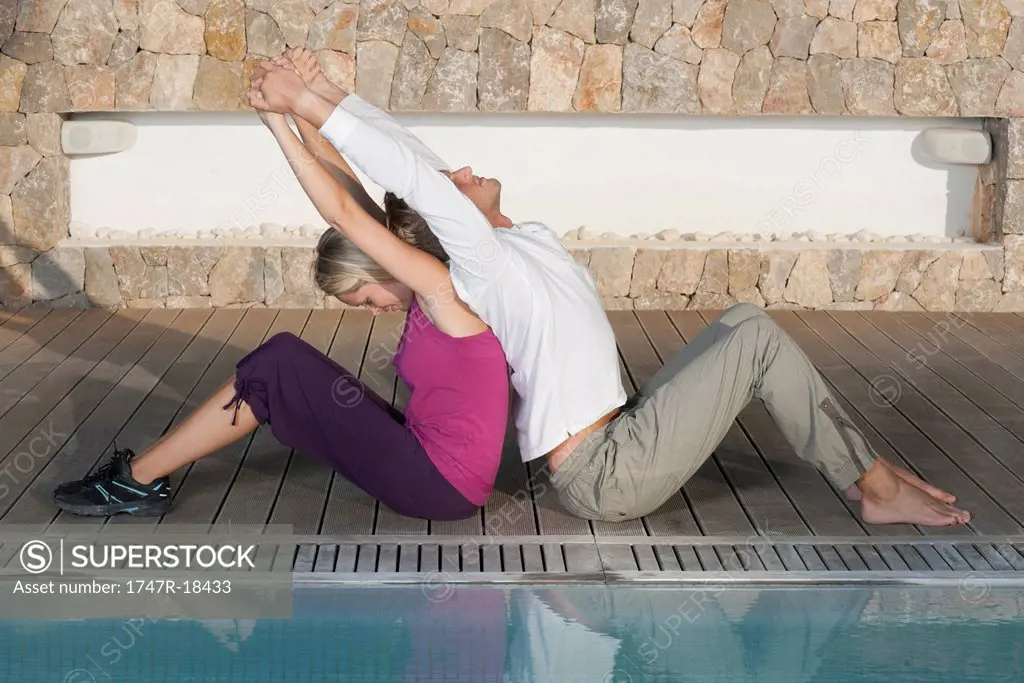 Young couple sitting back to back at poolside stretching