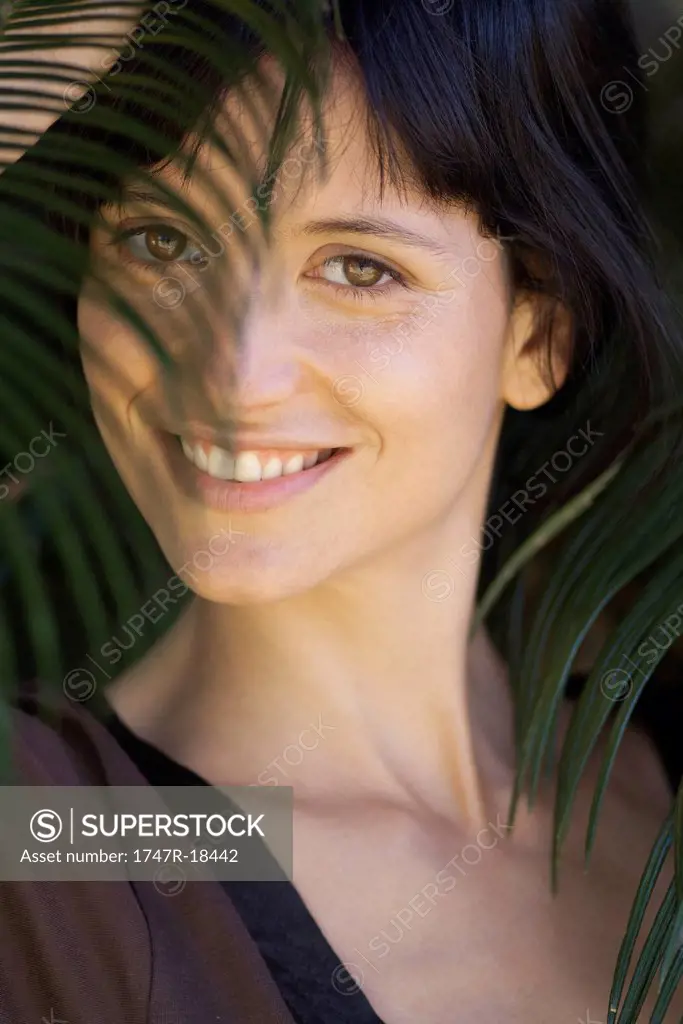 Young woman looking through palm leaf, smiling, portrait