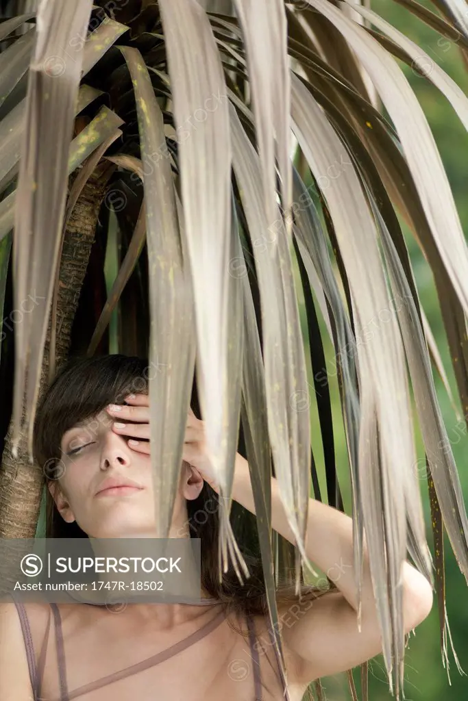 Young woman behind tropical foliage, hand covering one eye