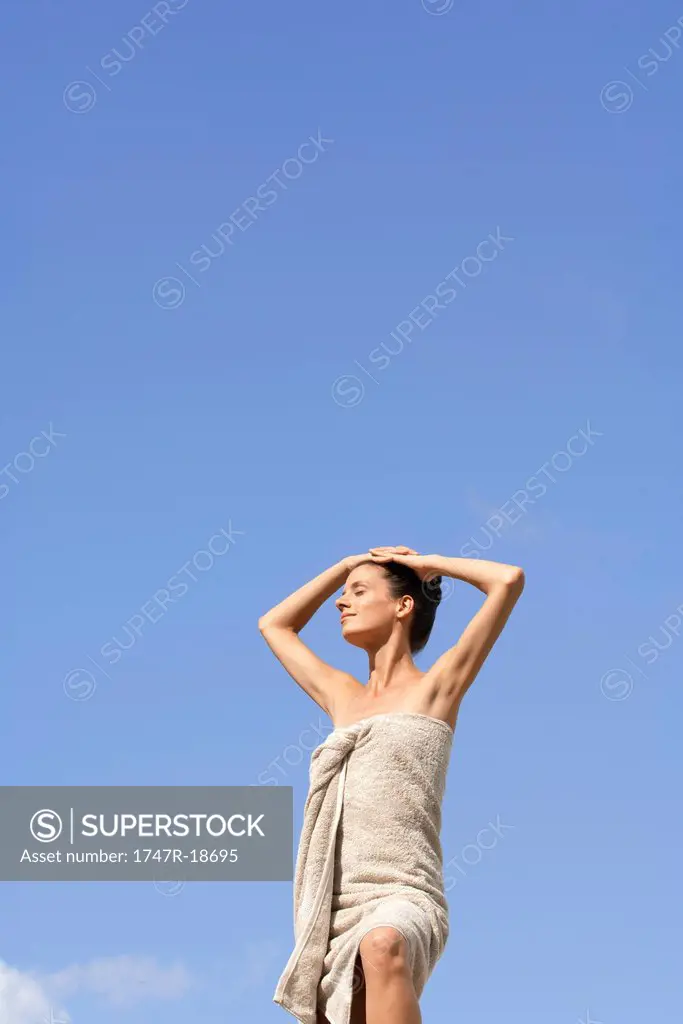 Mdi_adult woman standing wrapped in towel against blue sky, hands on head